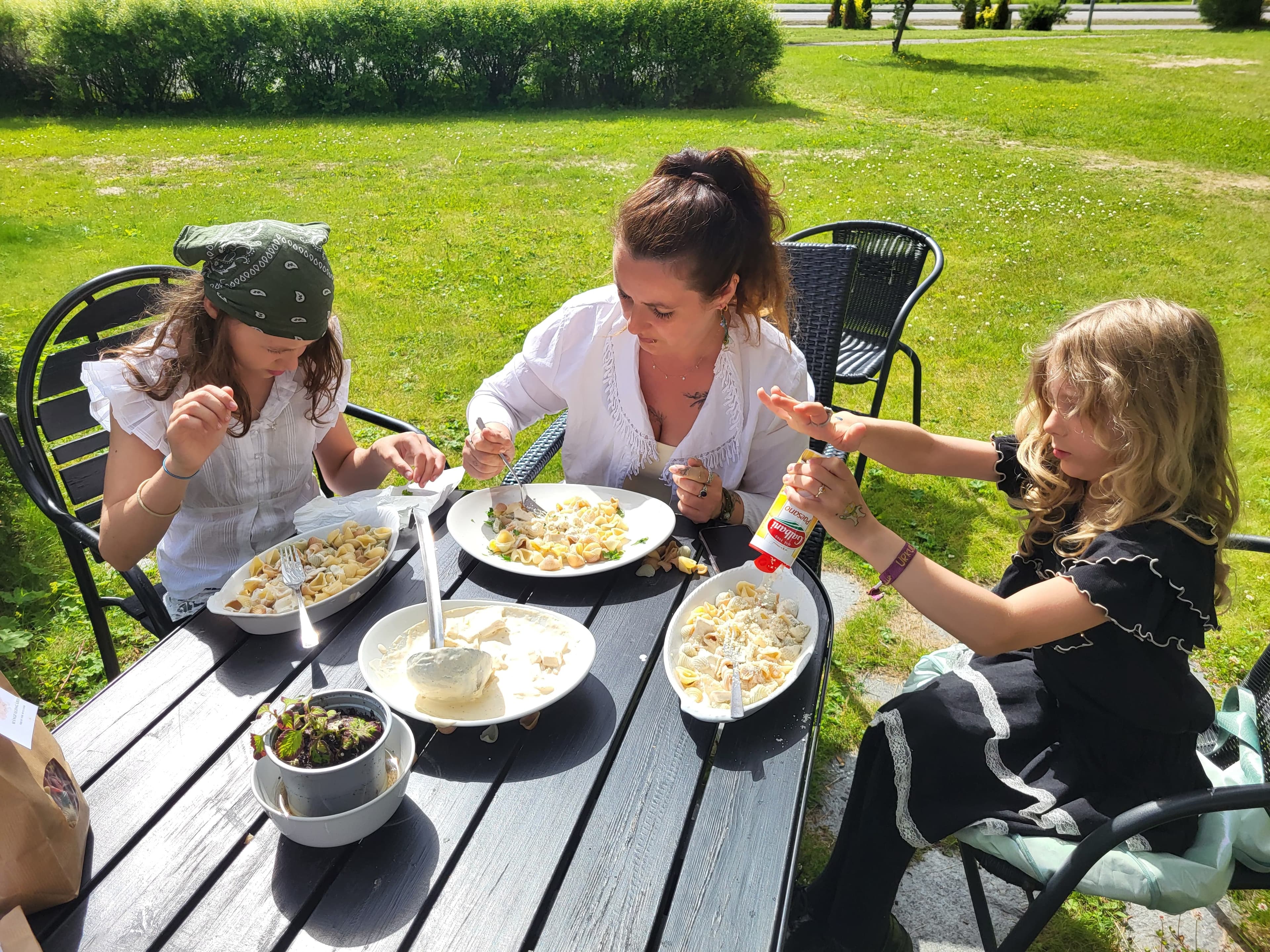 The family eating bowls of shell pasta together at a garden table in bright sunshine