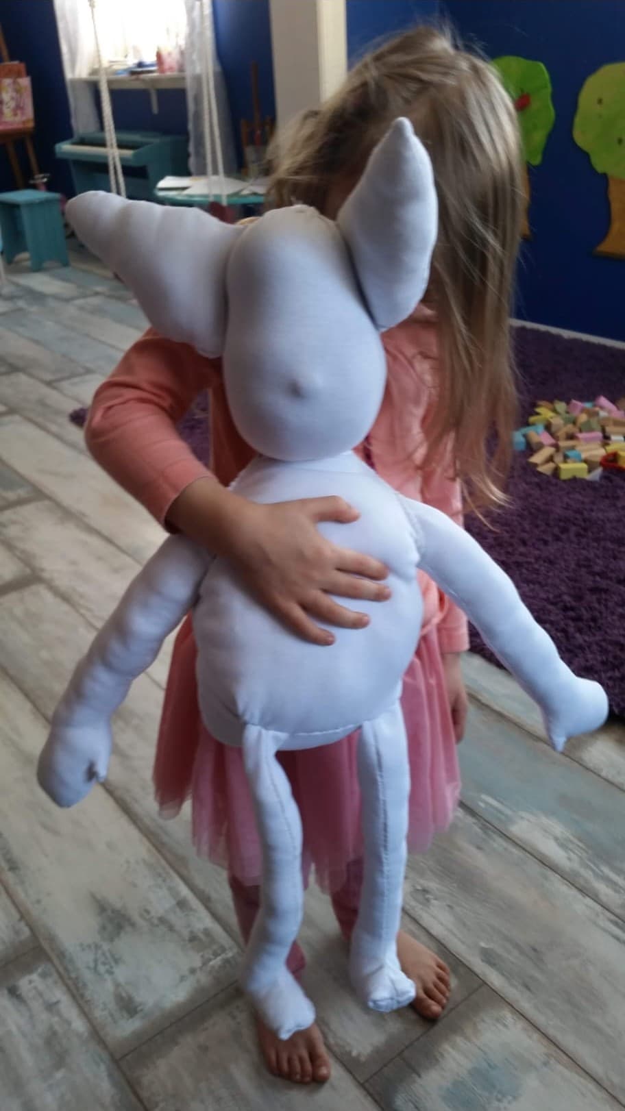 A small girl in a pink dress holding a large hand-sewn white doll with long ears, its face not yet made
