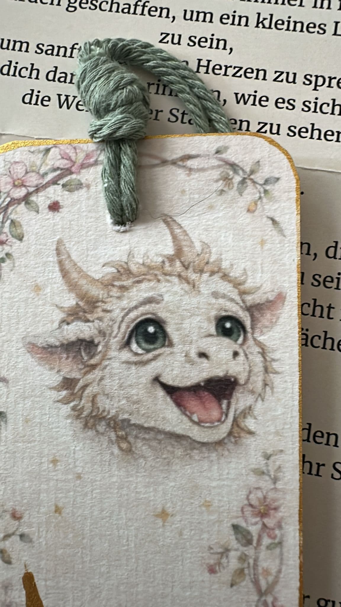 A gold-edged bookmark with Florenz's face and a green cord tassel, resting on an open book