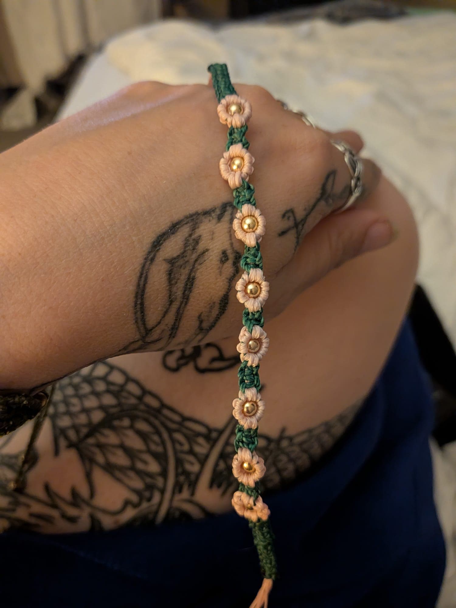 A green macramé bracelet with a row of peach flowers and gold beads, held over a hand