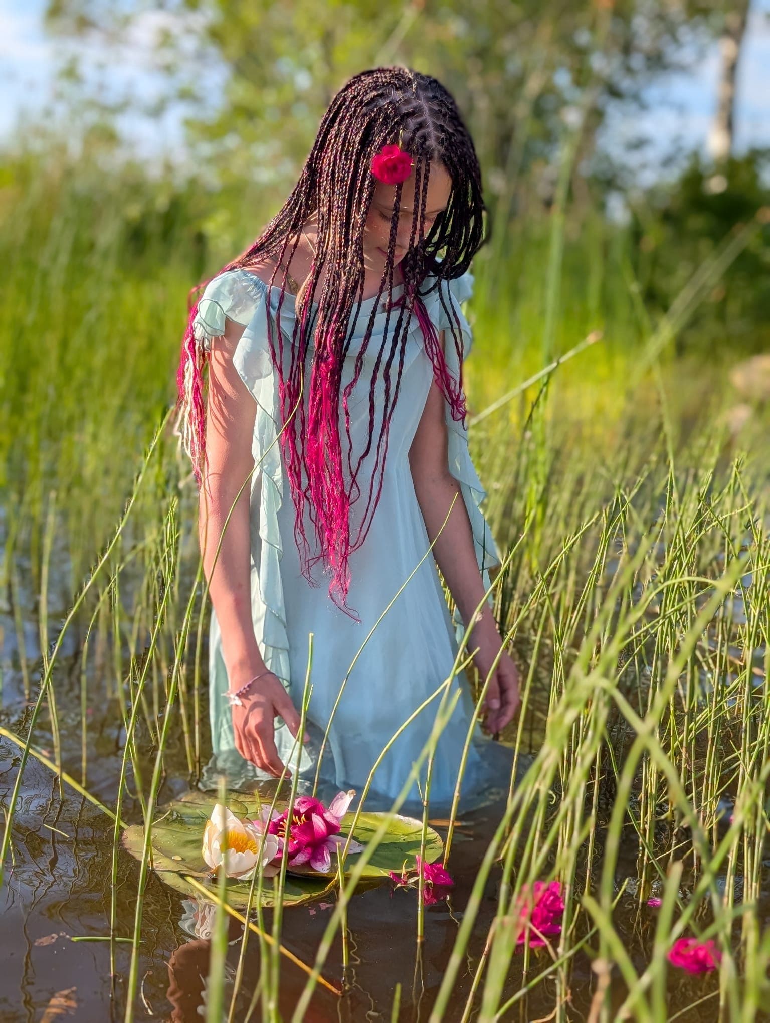 A girl with long braids and pink strands standing in the reeds, a red flower pinned above her ear