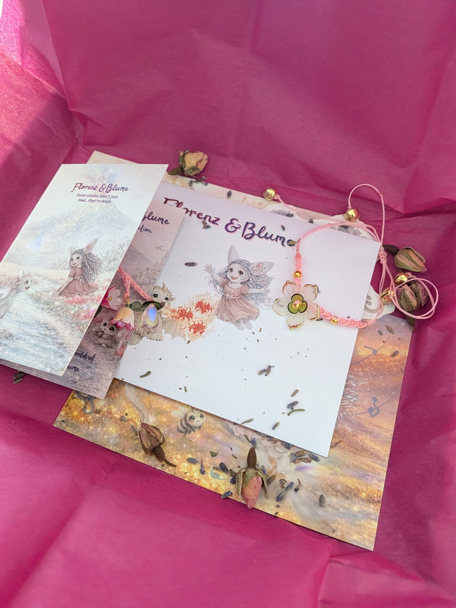 The set unwrapped on pink tissue: the booklets, a bookmark and a pink flower bracelet with little gold bells, scattered with dried rosebuds and lavender