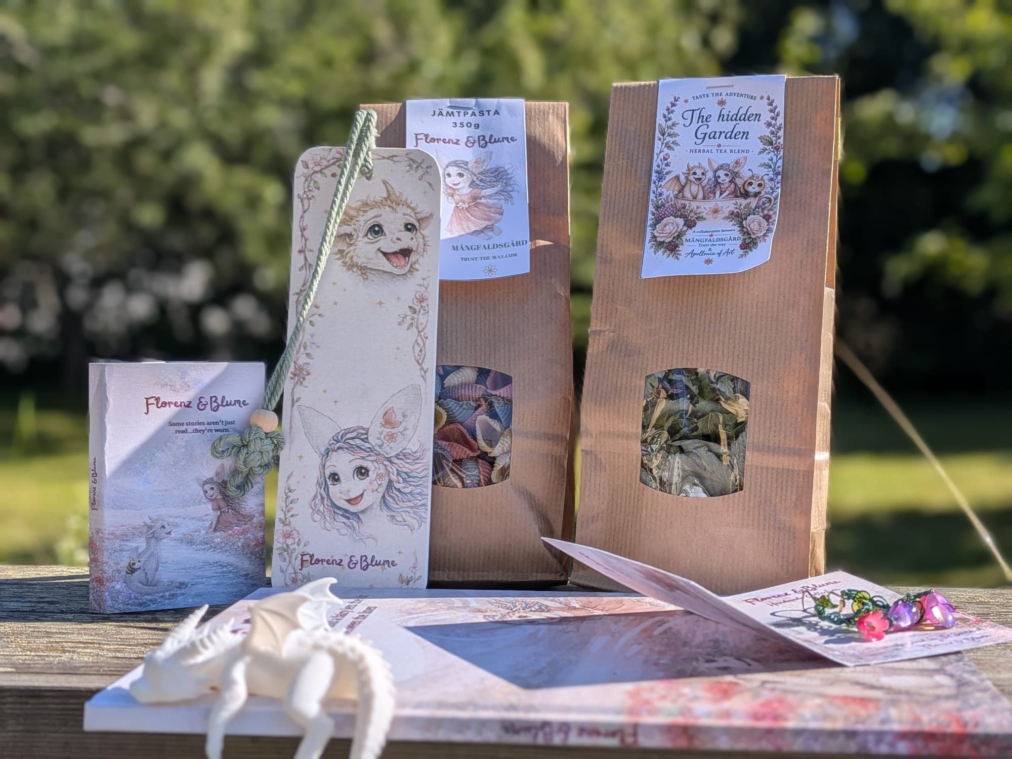 The Hidden Garden gift box laid out in the sun: the booklet, a bookmark, the pasta and tea bags, the dragon figure and a card of earrings