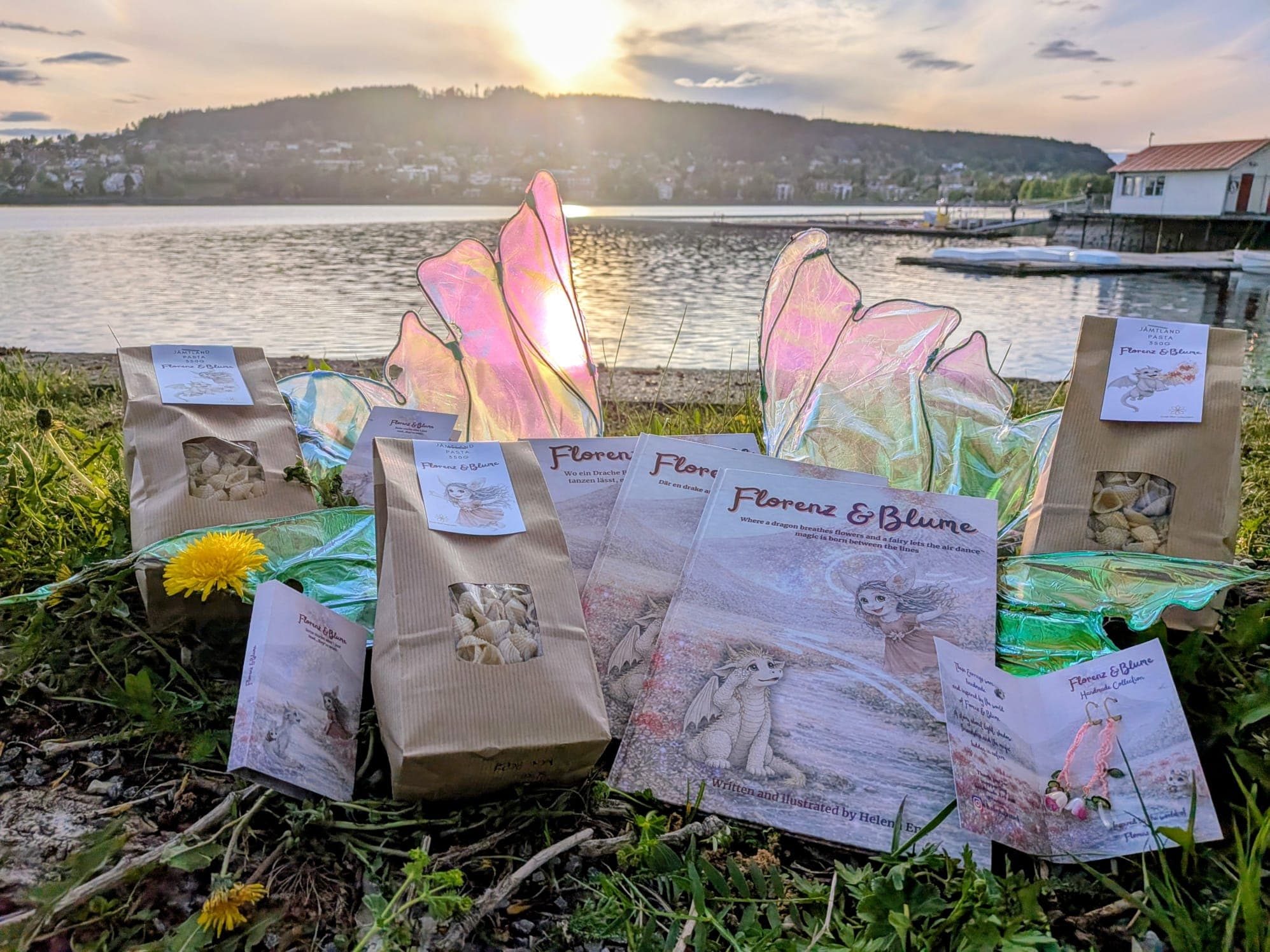 The whole range set out in the grass at the lakeside at sunset — books, pasta bags, fairy wings and cards