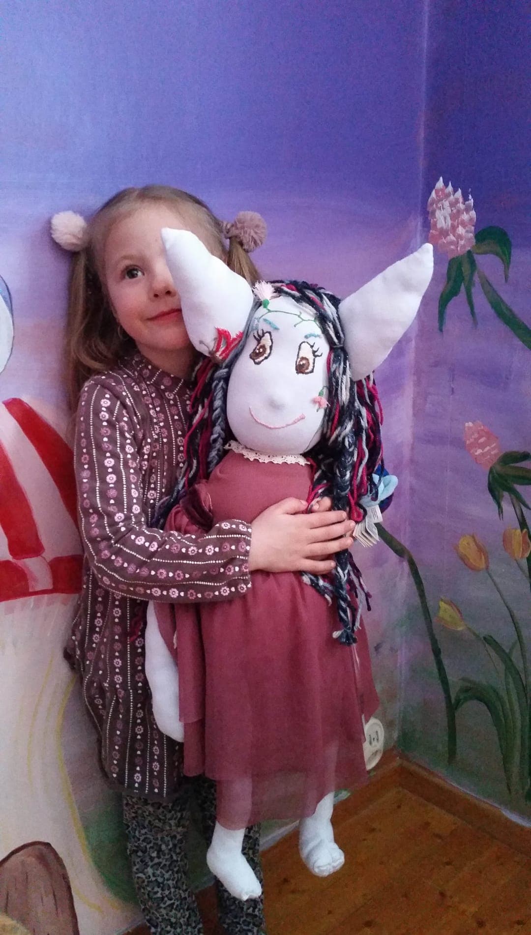 The same doll finished — embroidered eyes, braided yarn hair, a rose dress — hugged by a girl in front of a painted flower mural