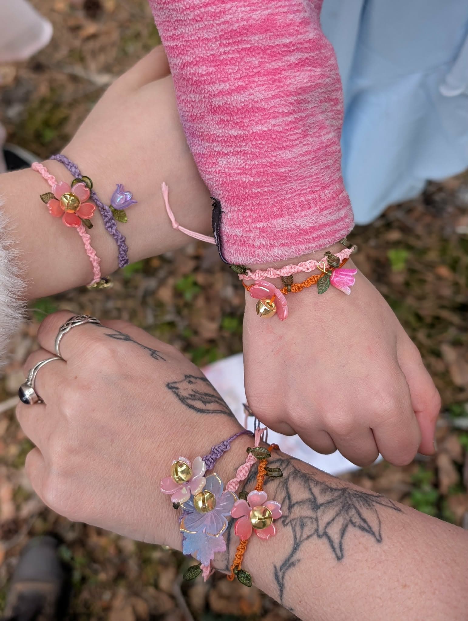 Three wrists held together over the forest floor, each wearing a knotted bracelet strung with resin flowers