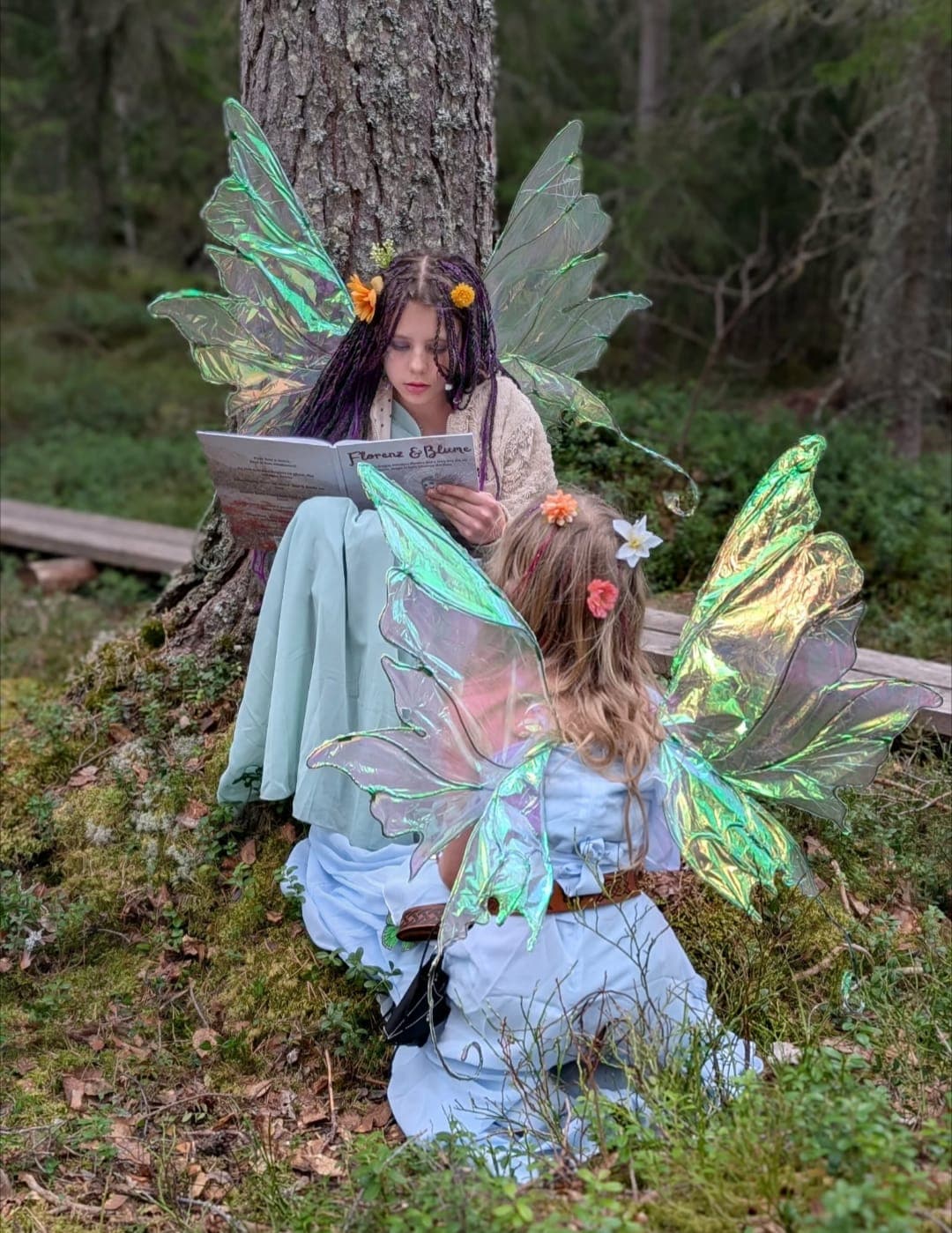 Two girls in iridescent fairy wings reading together against a pine tree